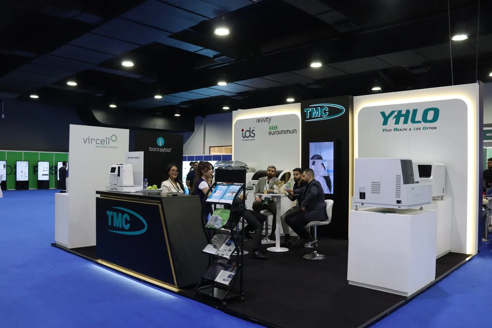 Wide view of the TMC exhibition stand showing TMC, Vircell, Bonraybio, Revvity IDS, EUROIMMUN and YHLO walls, with staff and visitors seated at a table