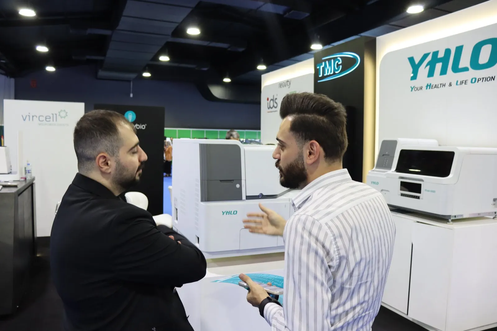 Staff member with a brochure talking to a visitor beside YHLO analysers under the YHLO sign