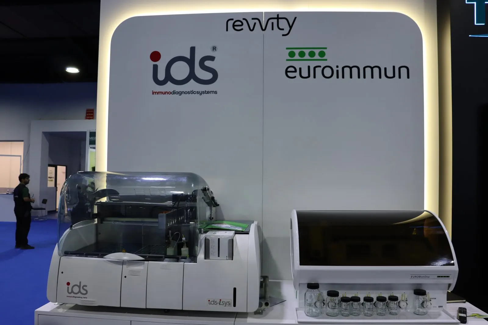 IDS-iSYS and EUROIMMUN analysers in front of the illuminated Revvity IDS and EUROIMMUN wall