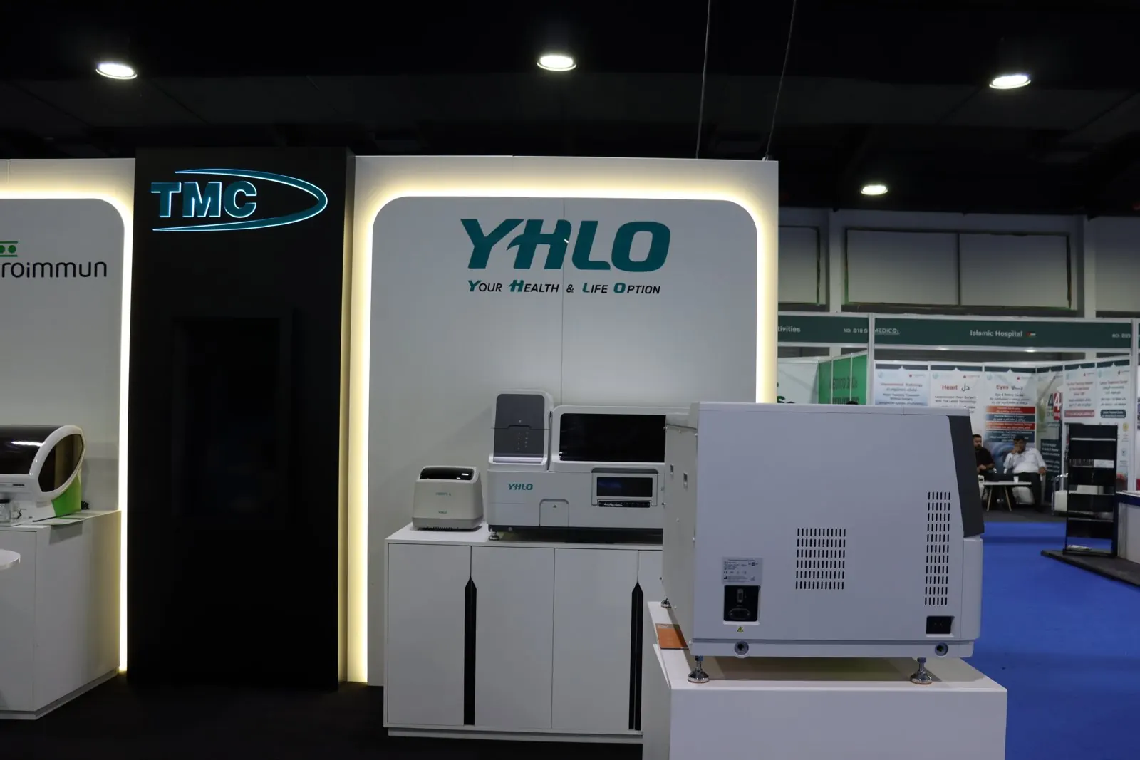 YHLO analysers on white cabinets in front of the illuminated YHLO wall beside the TMC logo