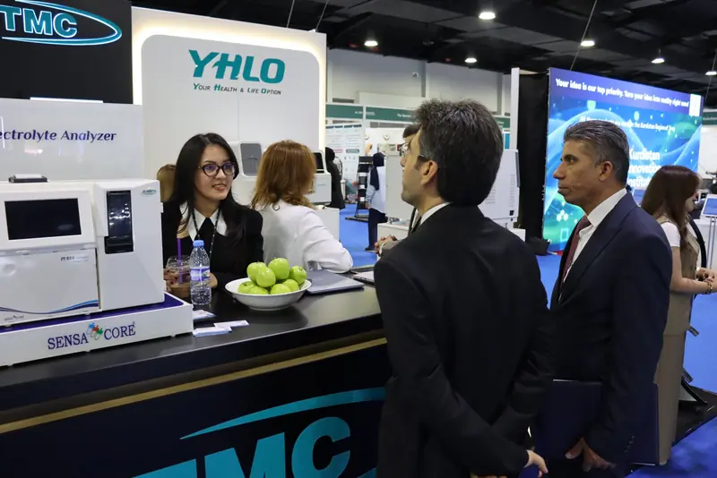 Staff member talking to two visitors across the TMC counter, with a Sensa Core ST-200 electrolyte analyser on display