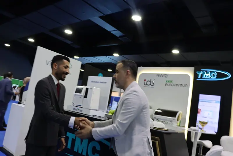 رجلان يتصافحان مبتسمين أمام منضدة TMC والجدار الإعلاني لـ Revvity IDS وEUROIMMUN