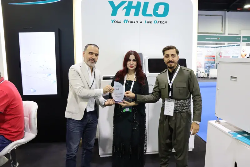 Three people holding a MEDICO plaque addressed to TMC Company in front of the YHLO wall