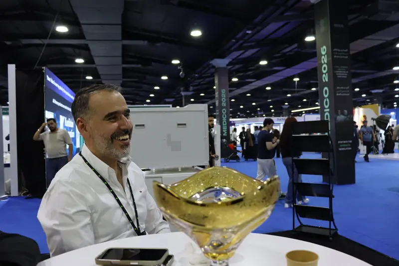 Smiling man seated at a table at the stand, with the exhibition hall and MEDICO 2026 pillar behind