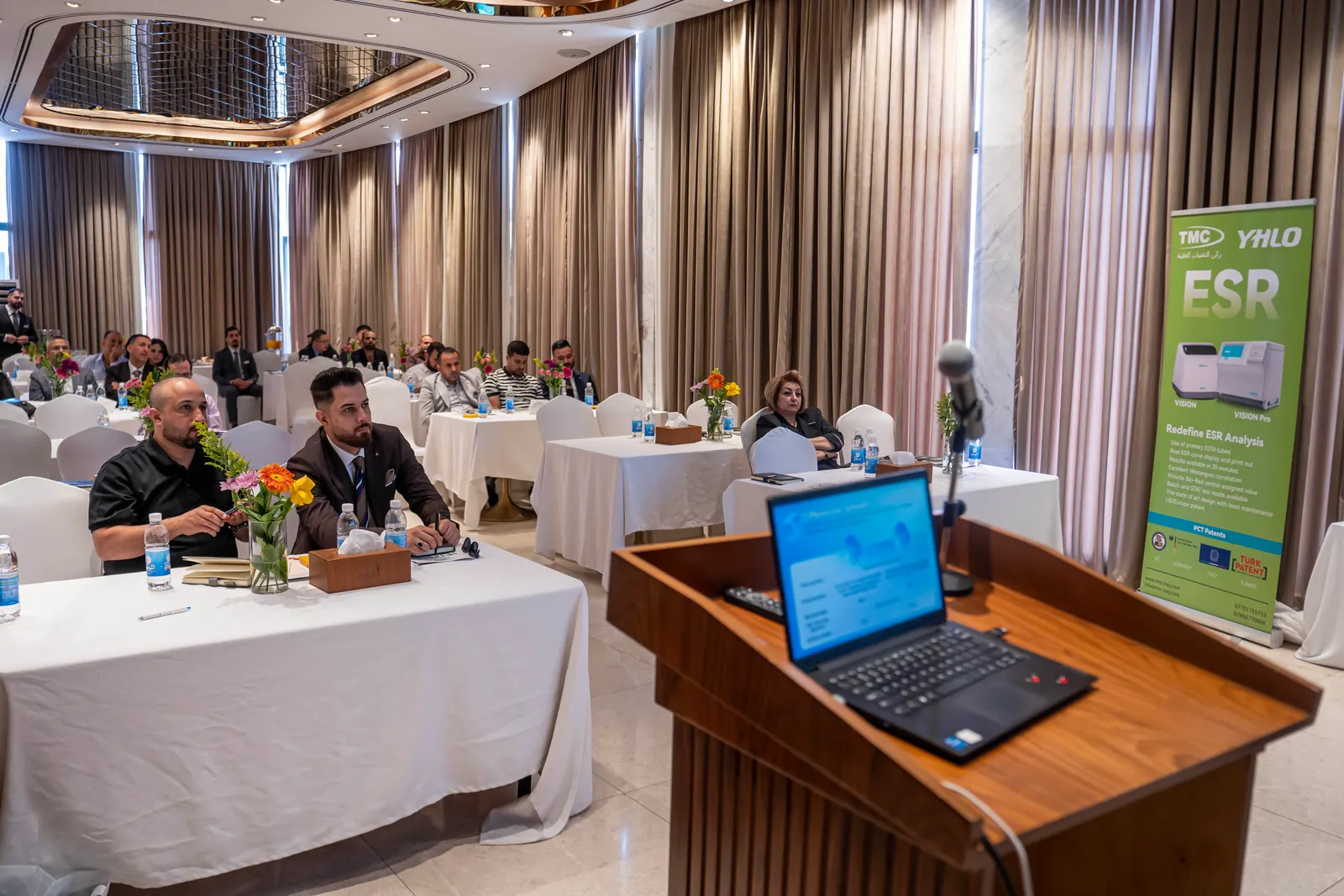 View from behind the podium laptop towards seated attendees, with a TMC and YHLO ESR banner on the right