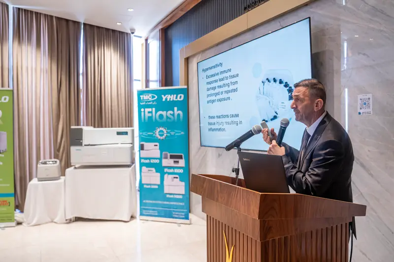 Speaker at a wooden podium presenting slides, with an iFlash banner and analysers to his left