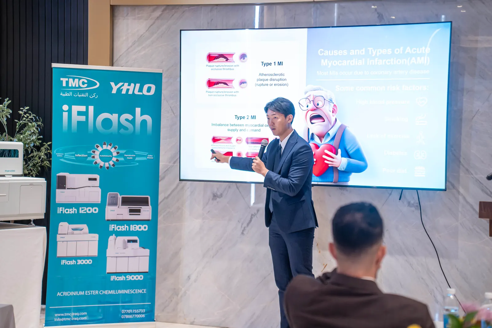 Speaker pointing to a slide on acute myocardial infarction, beside a TMC and YHLO iFlash banner